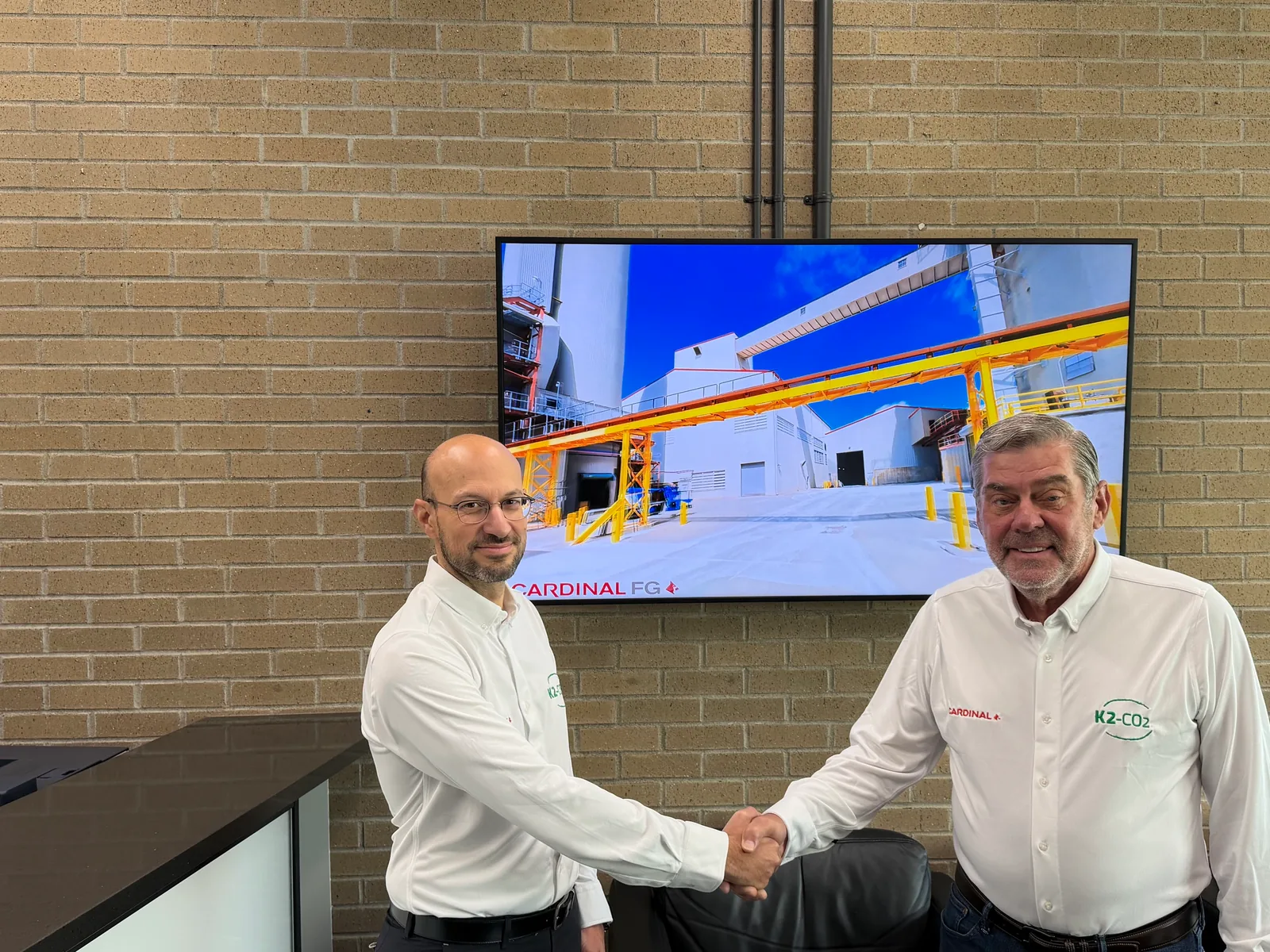 Alessandro Monteforte and Kelly Busch shake hands in front of a display showing a Cardinal FG float glass facility.